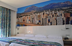 Habitació d'hotel amb un llit gran, una manta colorida, un mural amb un paisatge urbà a la paret i vistes a la muntanya.