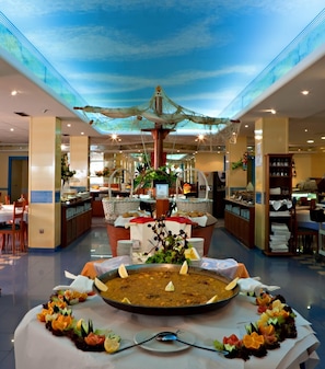 Un vestíbul d'hotel espaiós amb una zona de dinar central que destaca per una taula comuna gran, envoltada d'diverses zones de menjar i diferents disposicions de seients.