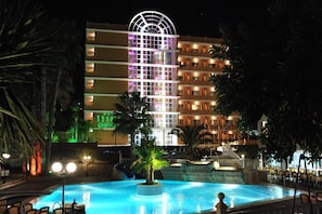 Un hotel amb piscina, palmeres i una façana d'edifici ben il·luminada.