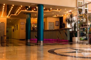 Vestíbulo de hotel con mostrador de recepción, pilares color turquesa, piso estampado y una partición de vidrio con una planta.