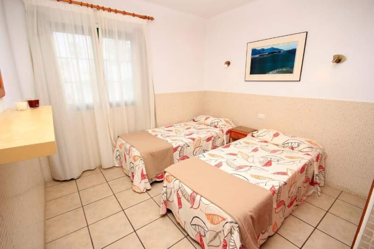Ein Zimmer mit zwei Betten, einem kleinen Tisch, einem Fenster mit transparenten Vorhängen und einem gerahmten Bild an der Wand.