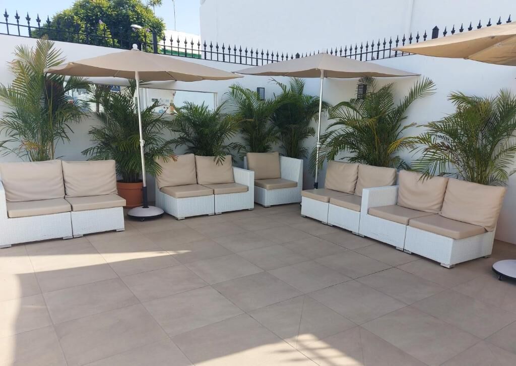 Een terras met witte wickerbanken en beige kussens, omringd door grote parasols en potpalmen.