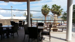 Un espace extérieur aménagé avec des tables et des chaises, offrant une vue sur une marina où sont amarrés des bateaux et où l’on peut admirer des palmiers.