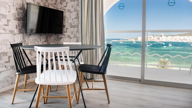 Une chambre moderne avec un mur en pierre, un téléviseur à écran plat, une table de salle à manger ronde avec quatre chaises et une grande fenêtre offrant une vue sur la mer et les bâtiments.