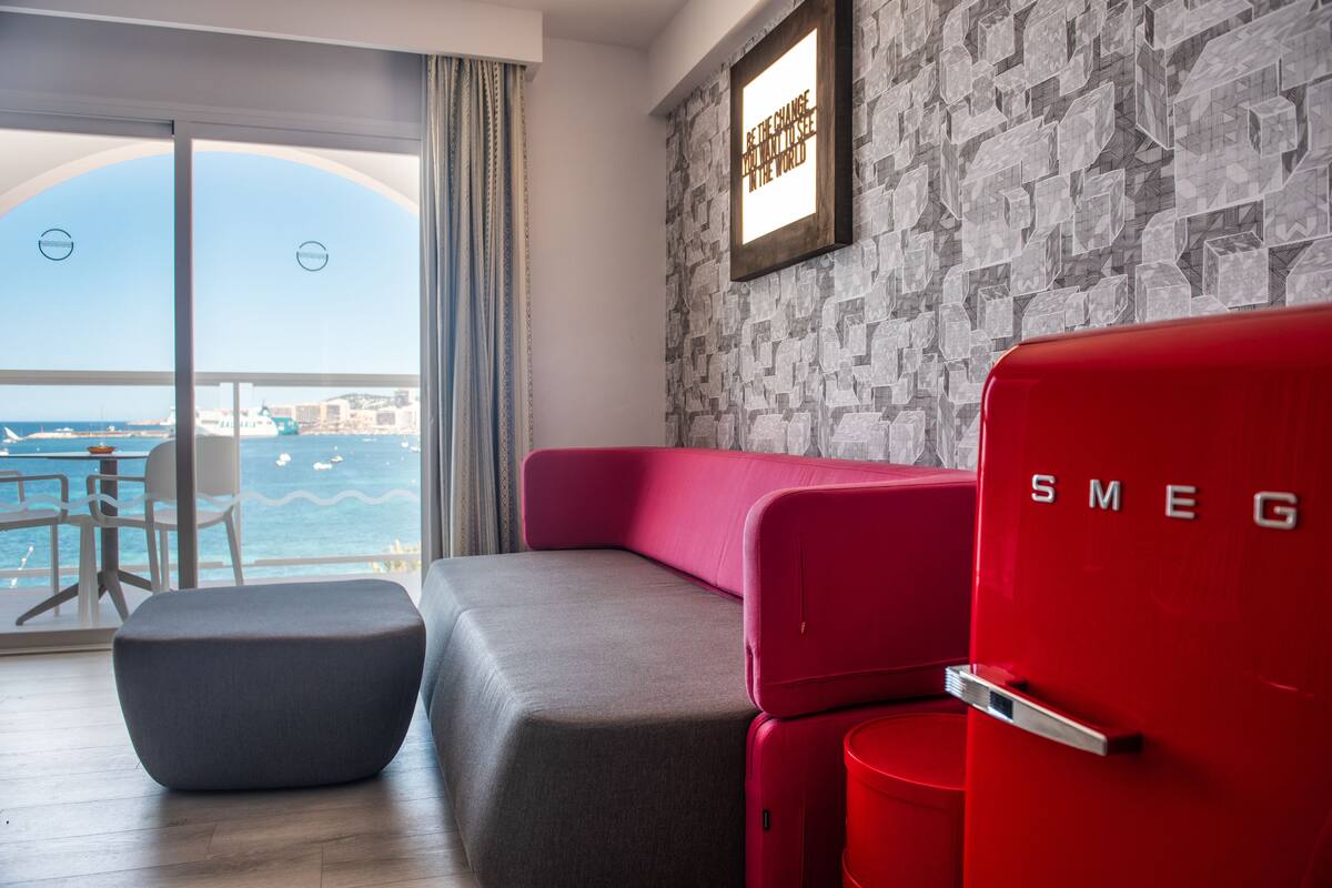 A modern room with a red and gray sofa, a small ottoman, and a red cabinet labeled "SMEG". A framed quote is on the wall, and the room has a view of the sea.