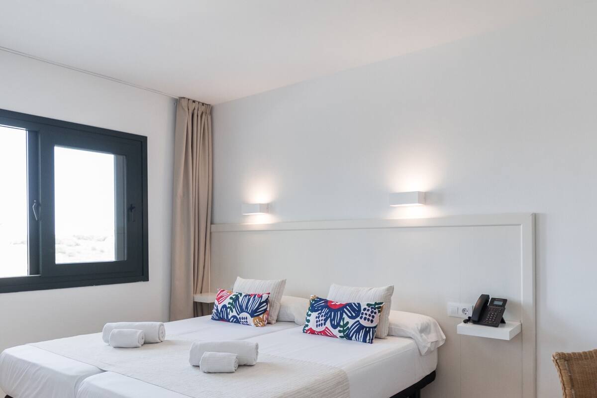A hotel room with two beds, a window with curtains, a bedside phone, and a wall-mounted light fixture.