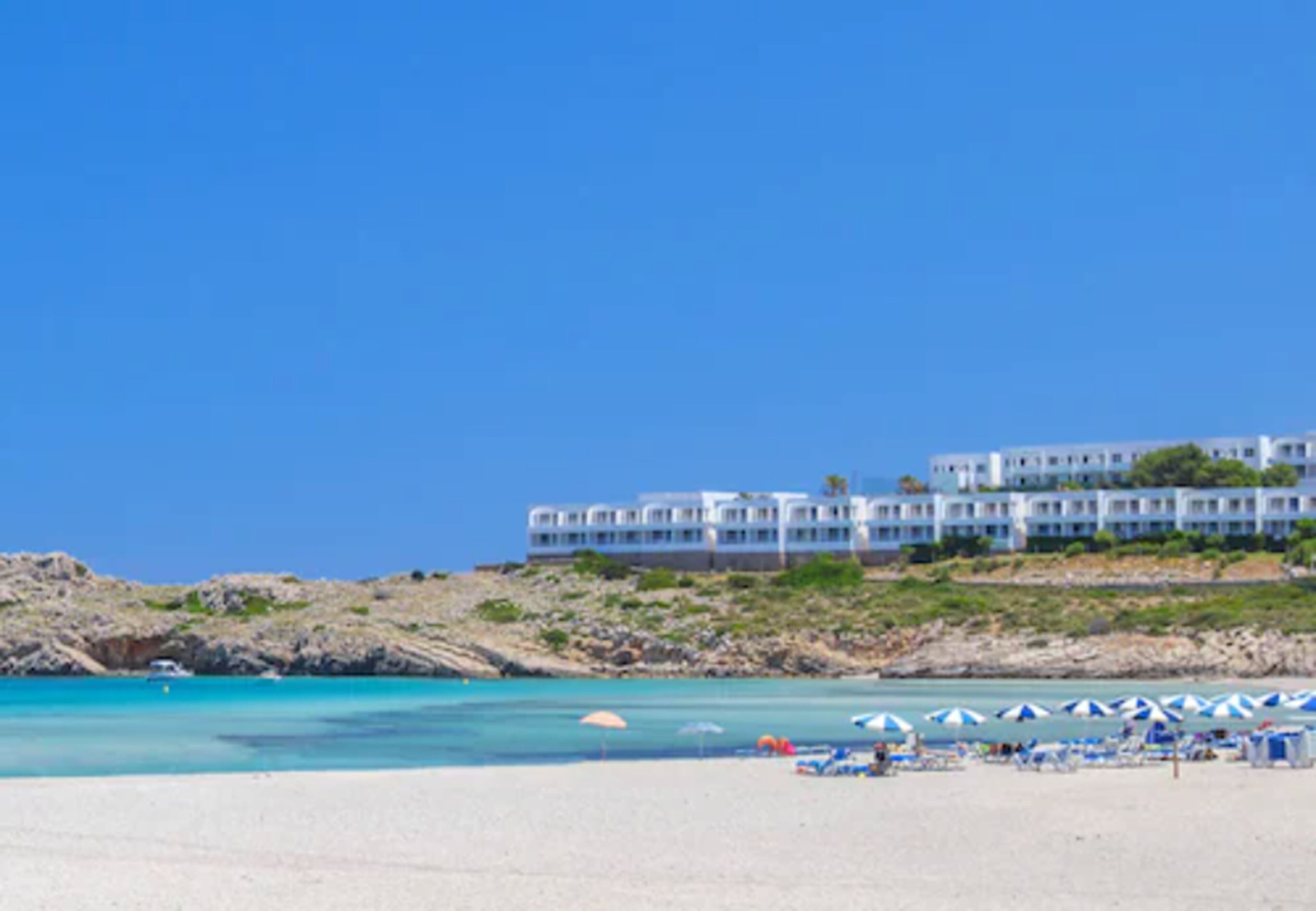 A beach with clear turquoise waters, a sandy shore, and a hotel with multiple floors and balconies overlooking the sea.