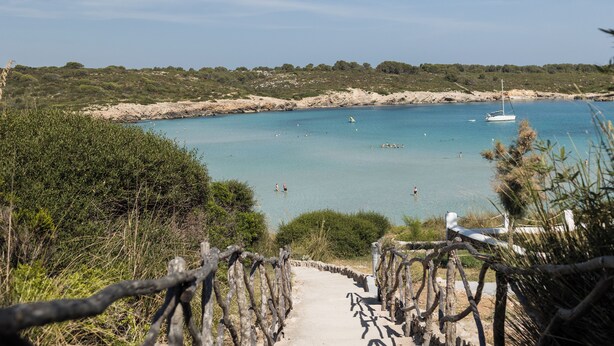 A coastal pathway leading to a beach with clear blue waters, a sailboat, and a rocky shoreline. - Aparthotel Beach Club (Mercadal)