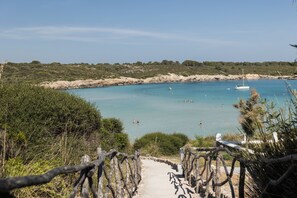 A coastal pathway leading to a beach with clear waters, boats, and sparse vegetation. - Aparthotel Beach Club (Mercadal)