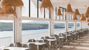 A dining area with tables and chairs, large windows with a view of the sea and hills, and hanging wicker lamps. - Aparthotel Beach Club (Mercadal)
