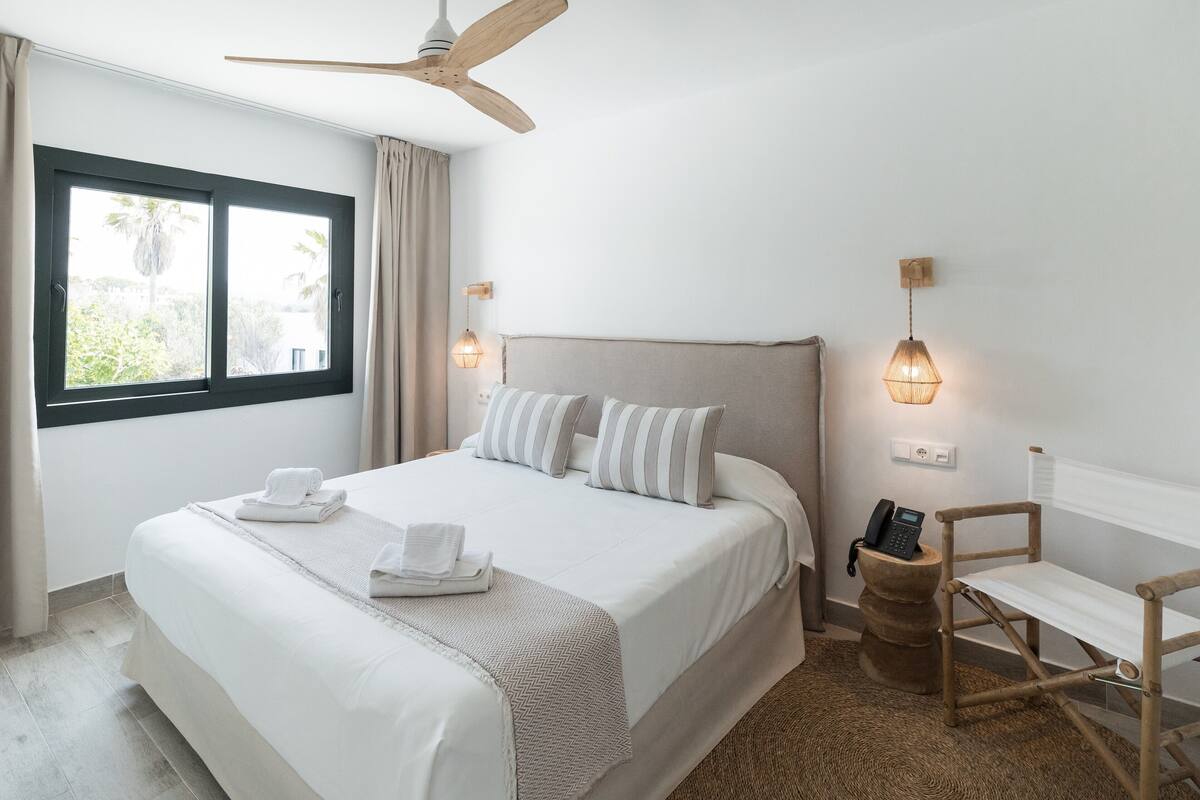 A neatly made bed with white linens and striped pillows, a wooden ceiling fan, a bedside table with a lamp, and a window with curtains.