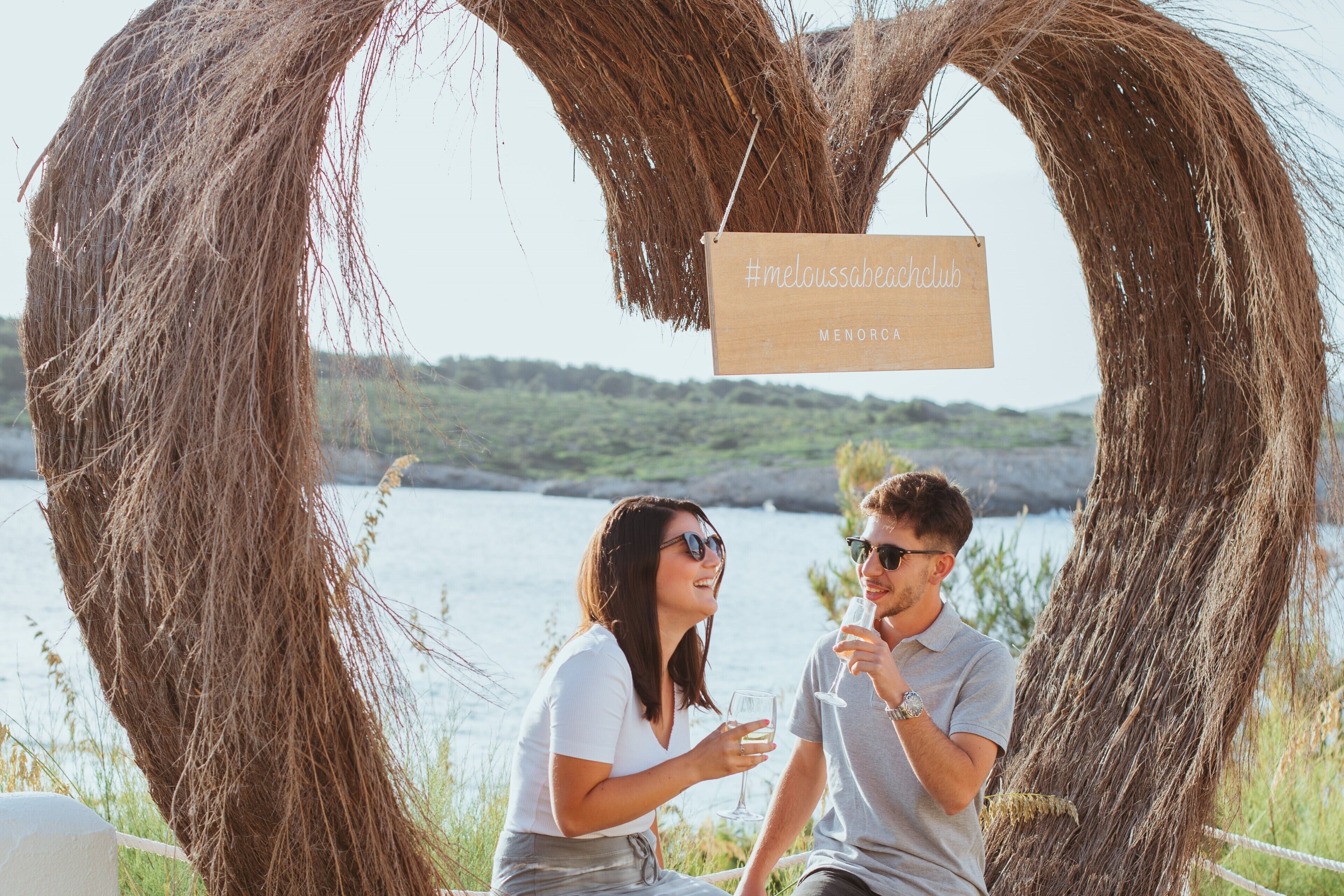 Un couple est assis sous une arche rustique en bois, sur laquelle on peut lire les inscriptions « meloussabeachclub » et « MENORQUE ».