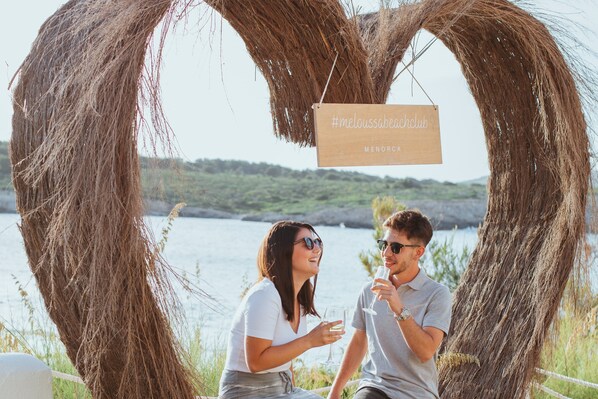 Una pareja sentada bajo un arco de madera rústico con un letrero que dice 'meloussabeachclub' y 'MENORCA'.