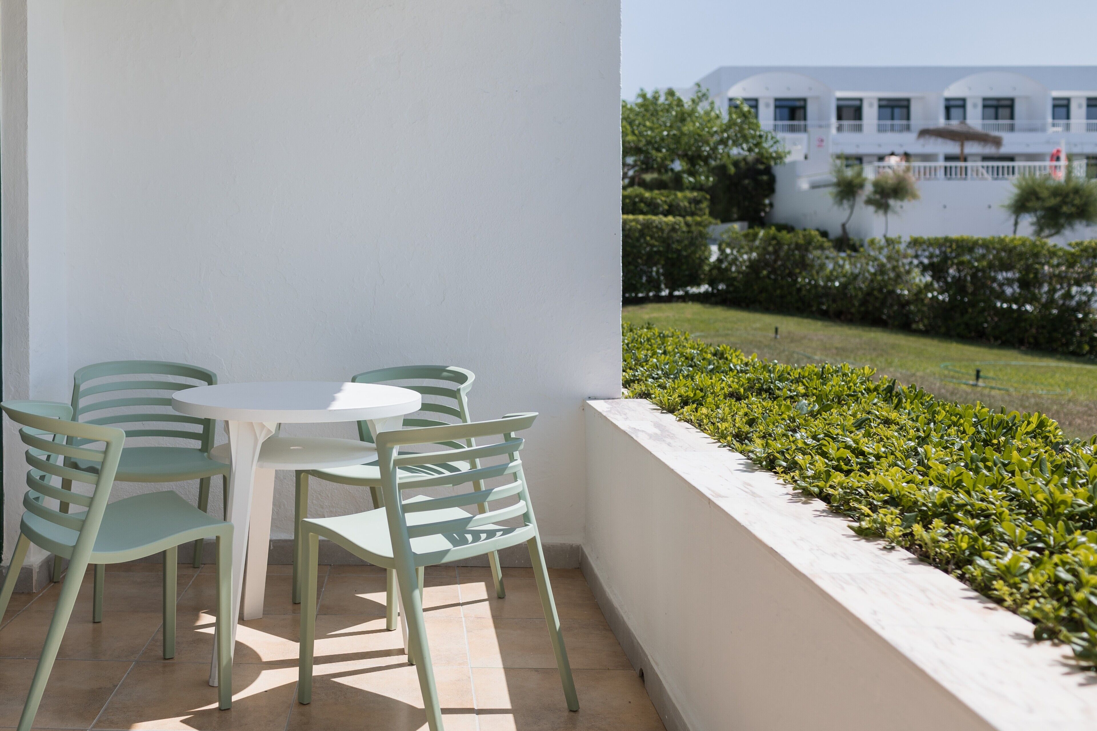 A balcony with a white table and four chairs, overlooking a well-maintained garden and a building with multiple windows.