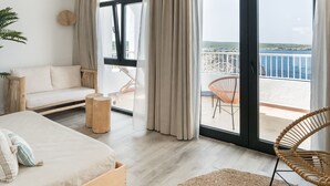A bedroom with a bed, a sofa, a wicker chair, and a balcony with a view of the sea. - Aparthotel Beach Club (Mercadal)
