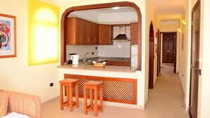 A compact kitchen with wooden cabinets, a built-in island with stools, and a bright yellow wall. - Callaomar (Adeje)