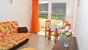 A room with a sofa, a table with a vase of flowers, and a view of mountains through a door. - Apartamentos Costamar (Arona)