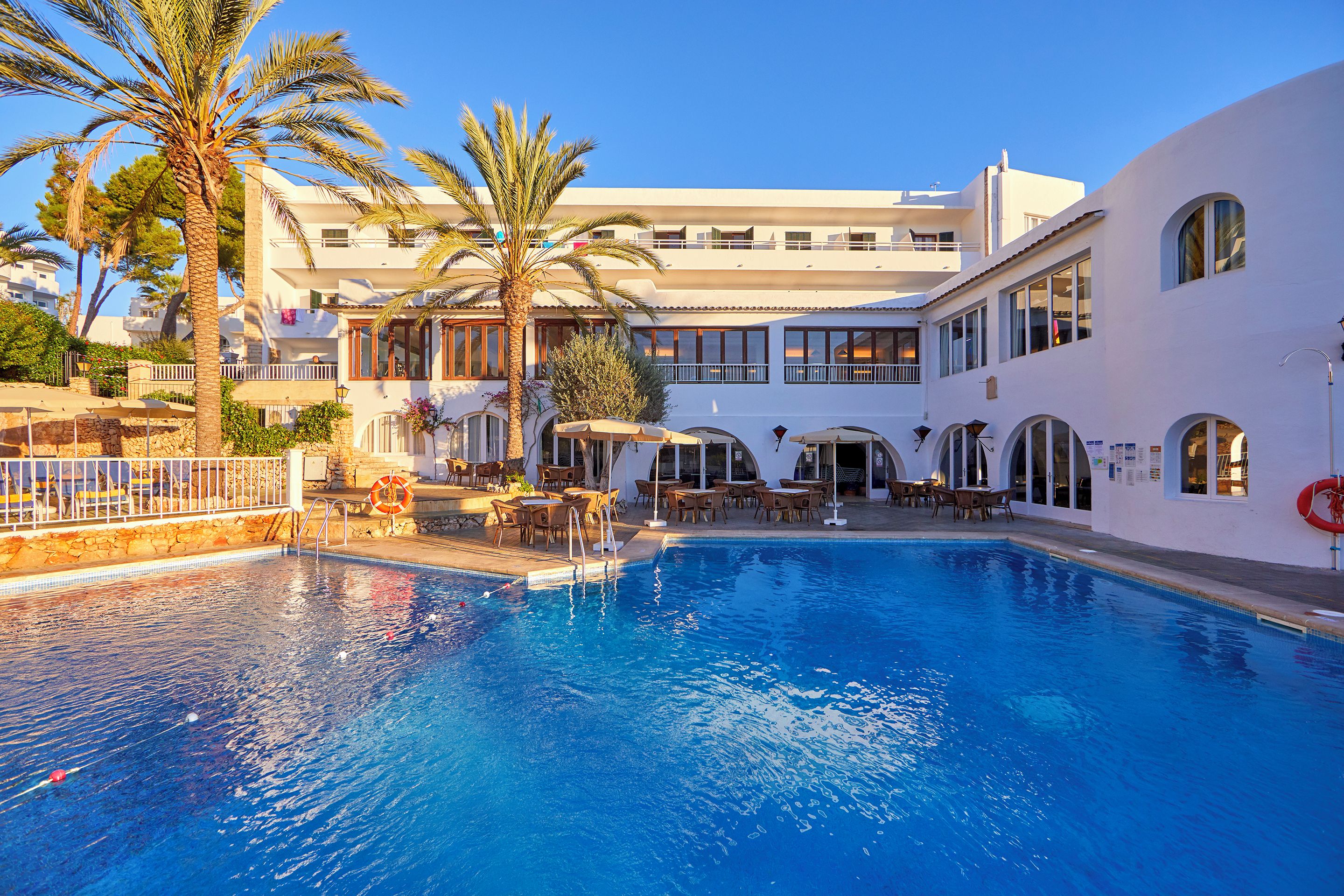 Une piscine entourée de chaises longues et de parasols, le tout bordé par un bâtiment doté de nombreux balcons et de fenêtres.