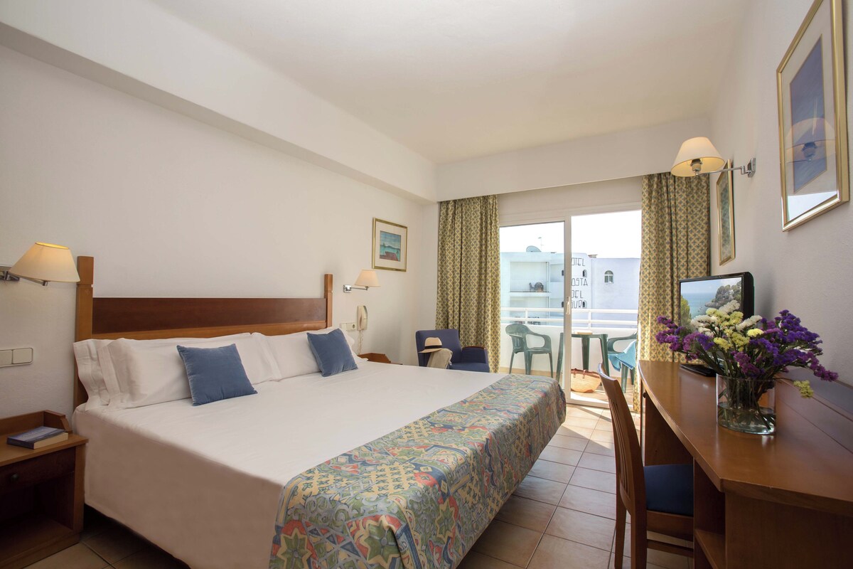Une chambre d’hôtel avec un lit, un bureau, une chaise, une lampe, un vase avec des fleurs et une fenêtre avec des rideaux.