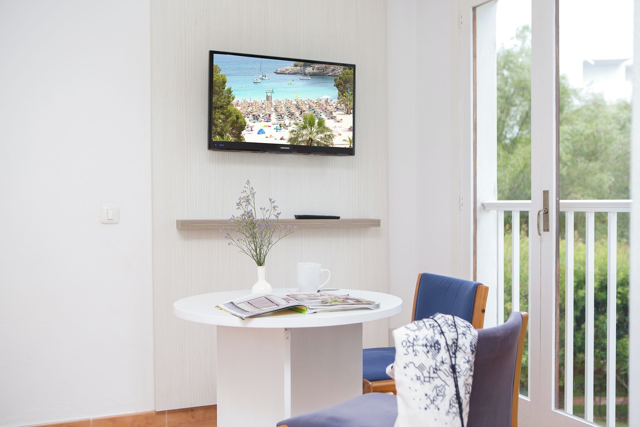 Ein Zimmer mit an der Wand befestigtem Fernseher, der eine Strandansicht zeigt, einem kleinen runden Tisch mit einer Vase und Zeitschriften, einem blauen Stuhl und Blick ins Grüne durch eine Schiebetür.