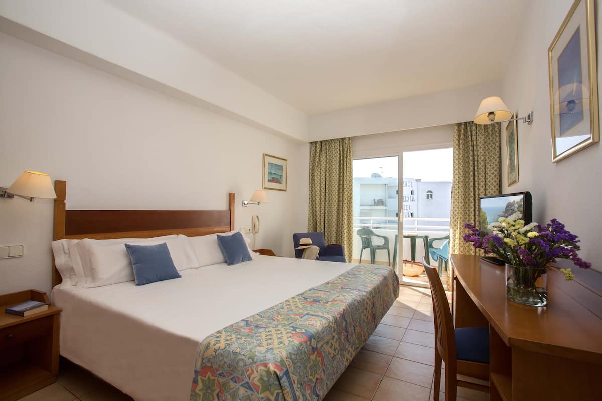 Une chambre d’hôtel avec un lit, un bureau, une chaise, une lampe, un vase avec des fleurs et une fenêtre avec des rideaux.