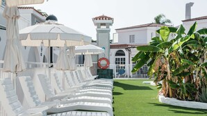 A row of white lounge chairs with umbrellas, a green lawn, and a building with a tower in the background. - Bungalows Parque Nogal (San Bartolomé de Tirajana)