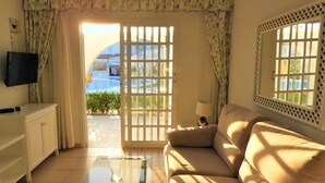 A living room with a sofa, a TV, a glass coffee table, and a view of a pool and greenery outside. - Bungalows Parque Nogal (San Bartolomé de Tirajana)