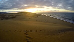 A beach with footprints leading to the water, a sunset in the background, and a cloudy sky. - Bungalows Parque Nogal (San Bartolomé de Tirajana)