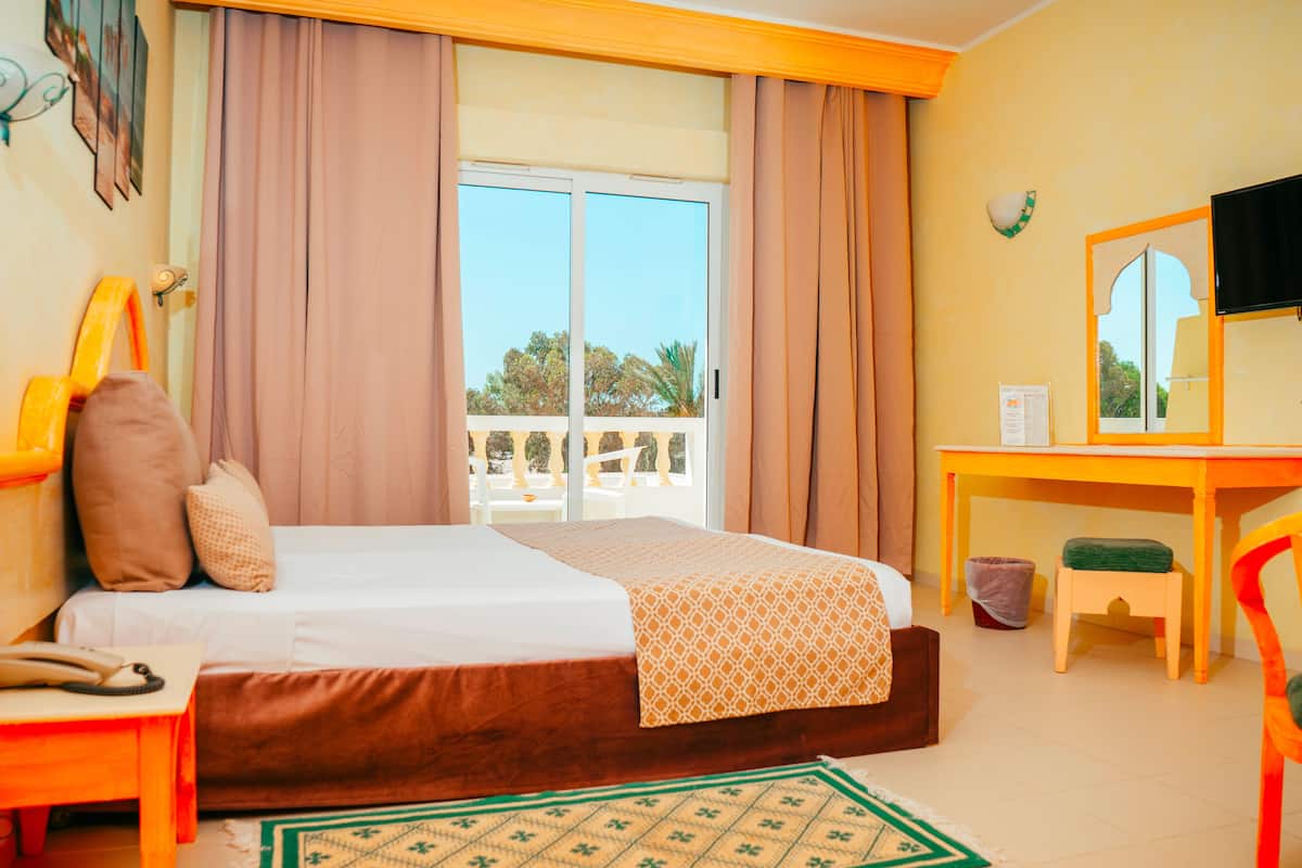 Habitación de hotel con cama, escritorio, silla, taburete pequeño, teléfono y ventana con cortinas.