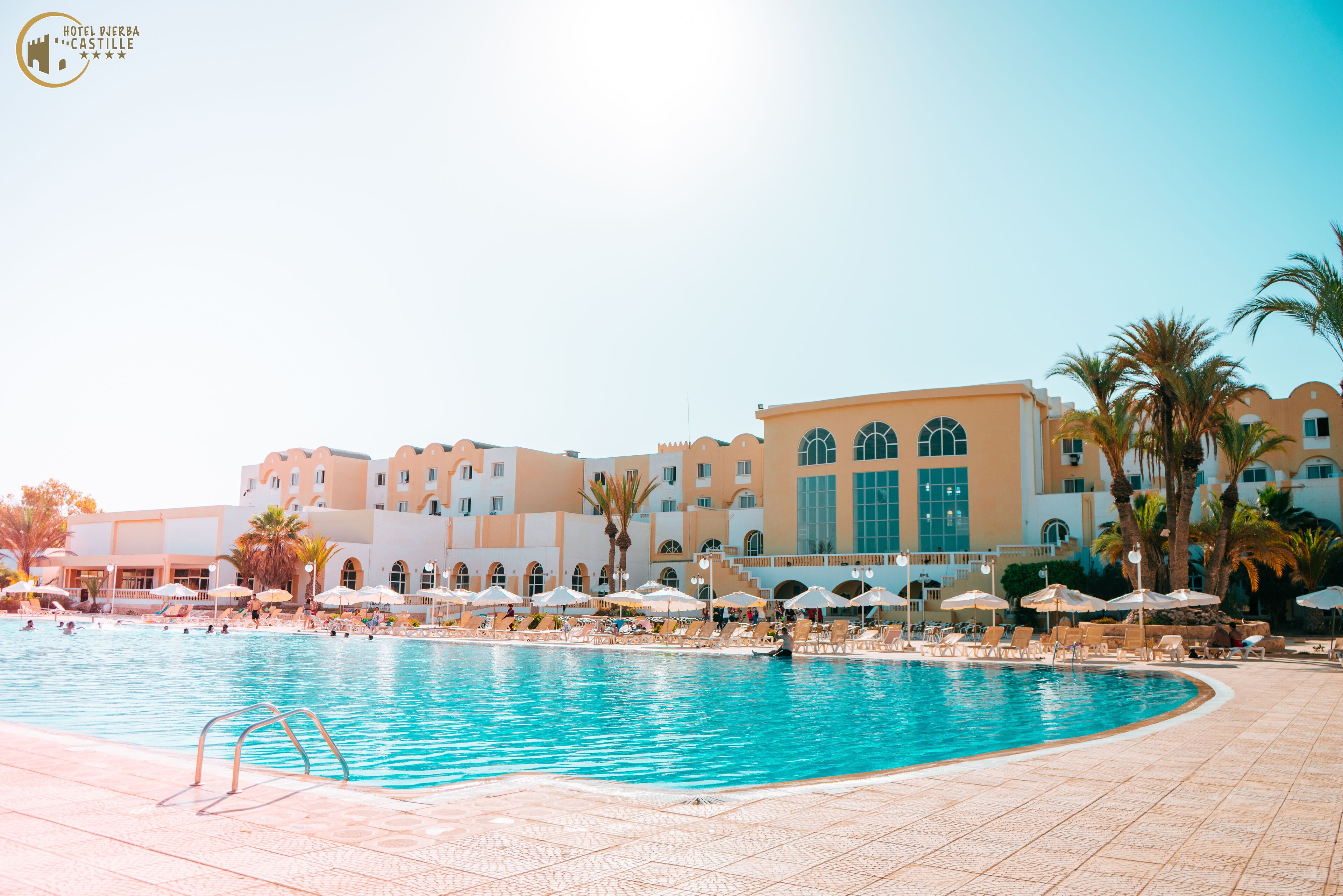 Uma área de piscina de hotel com várias espreguiçadeiras e guarda-sóis, cercada por palmeiras e um edifício grande com janelas em arco.