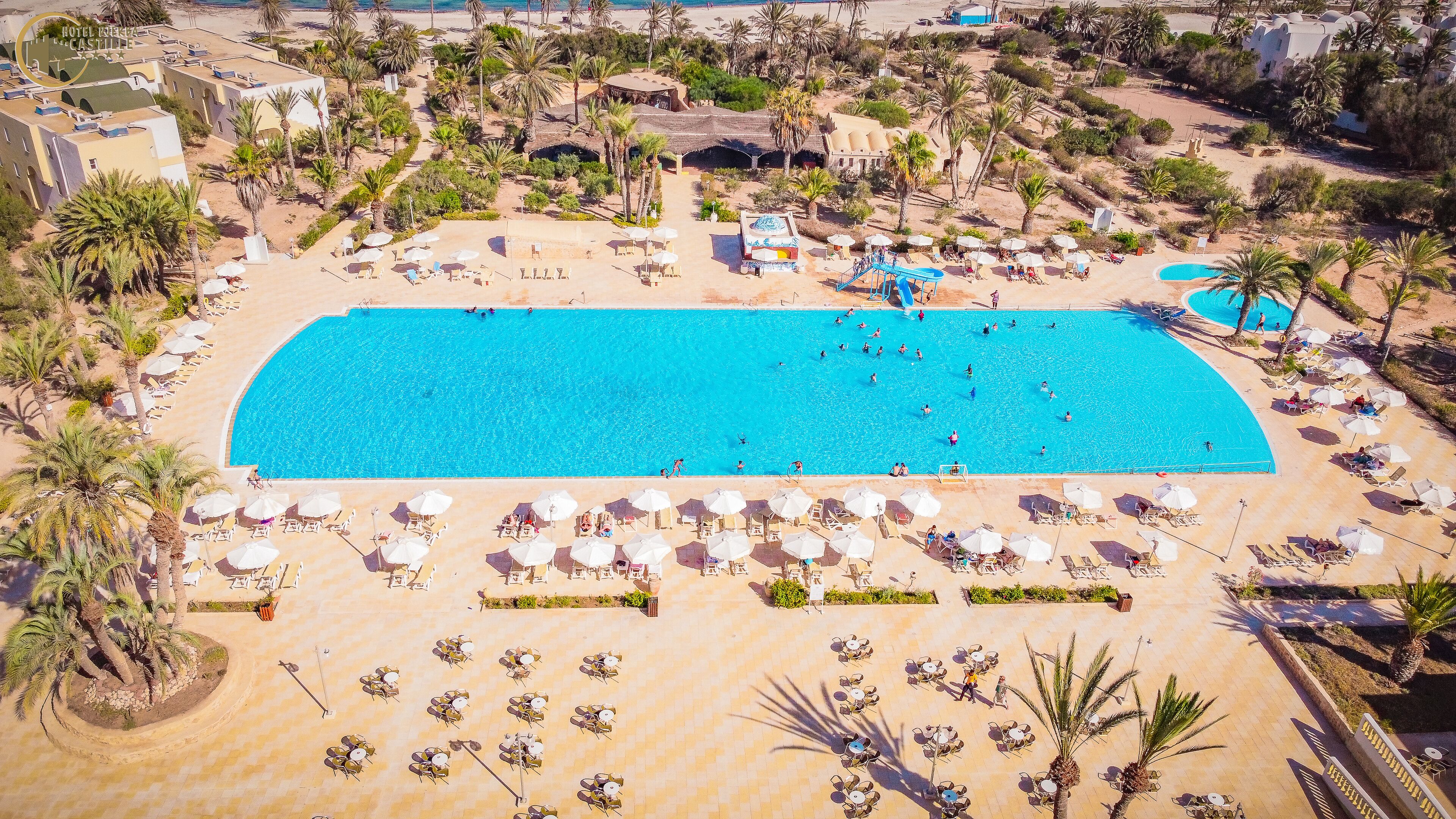 Una grande piscina circondata da lettini e ombrelloni, con palme e un paesaggio desertico sullo sfondo.