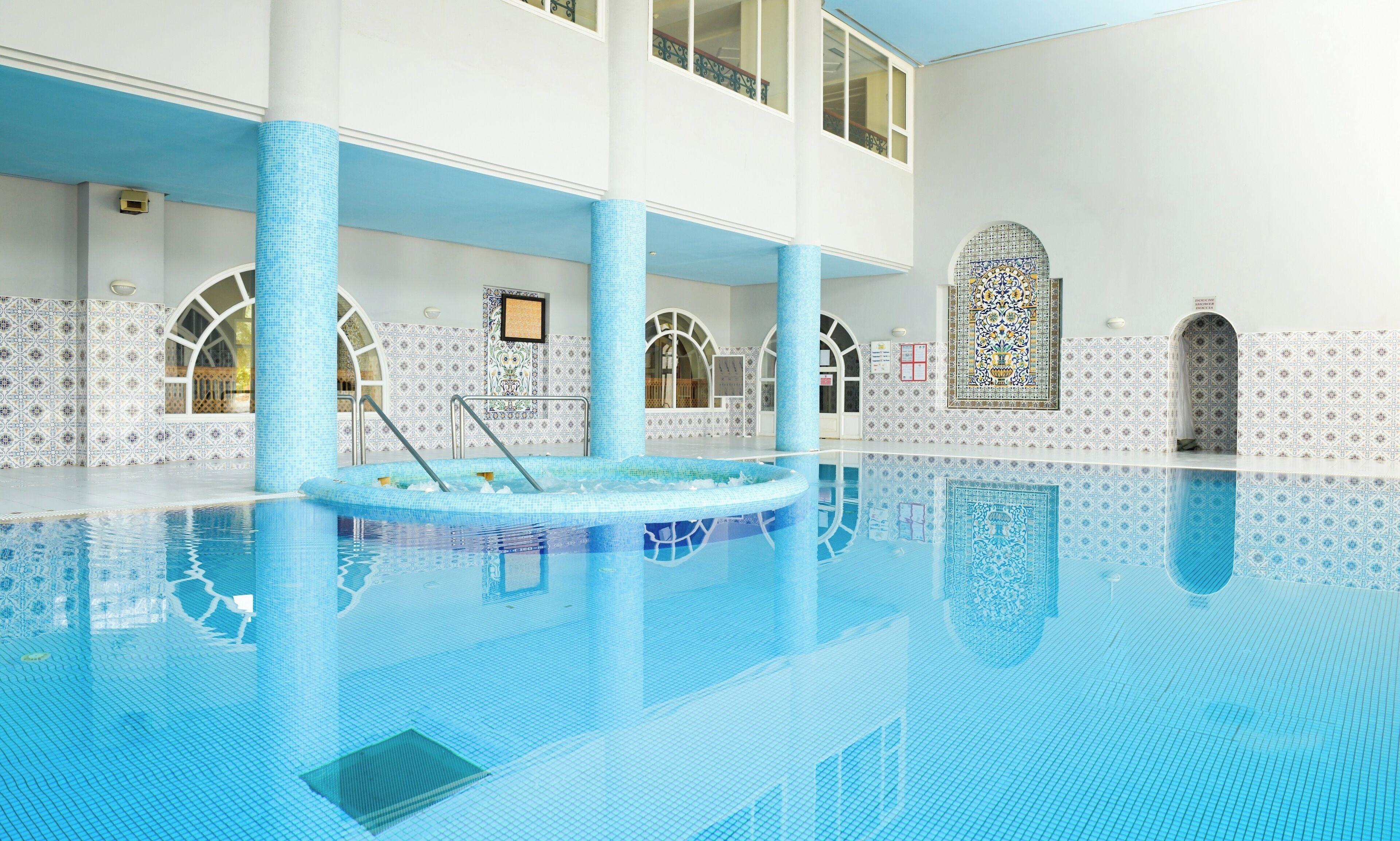 A large indoor swimming pool with a tiled floor and walls, surrounded by white columns and arches.