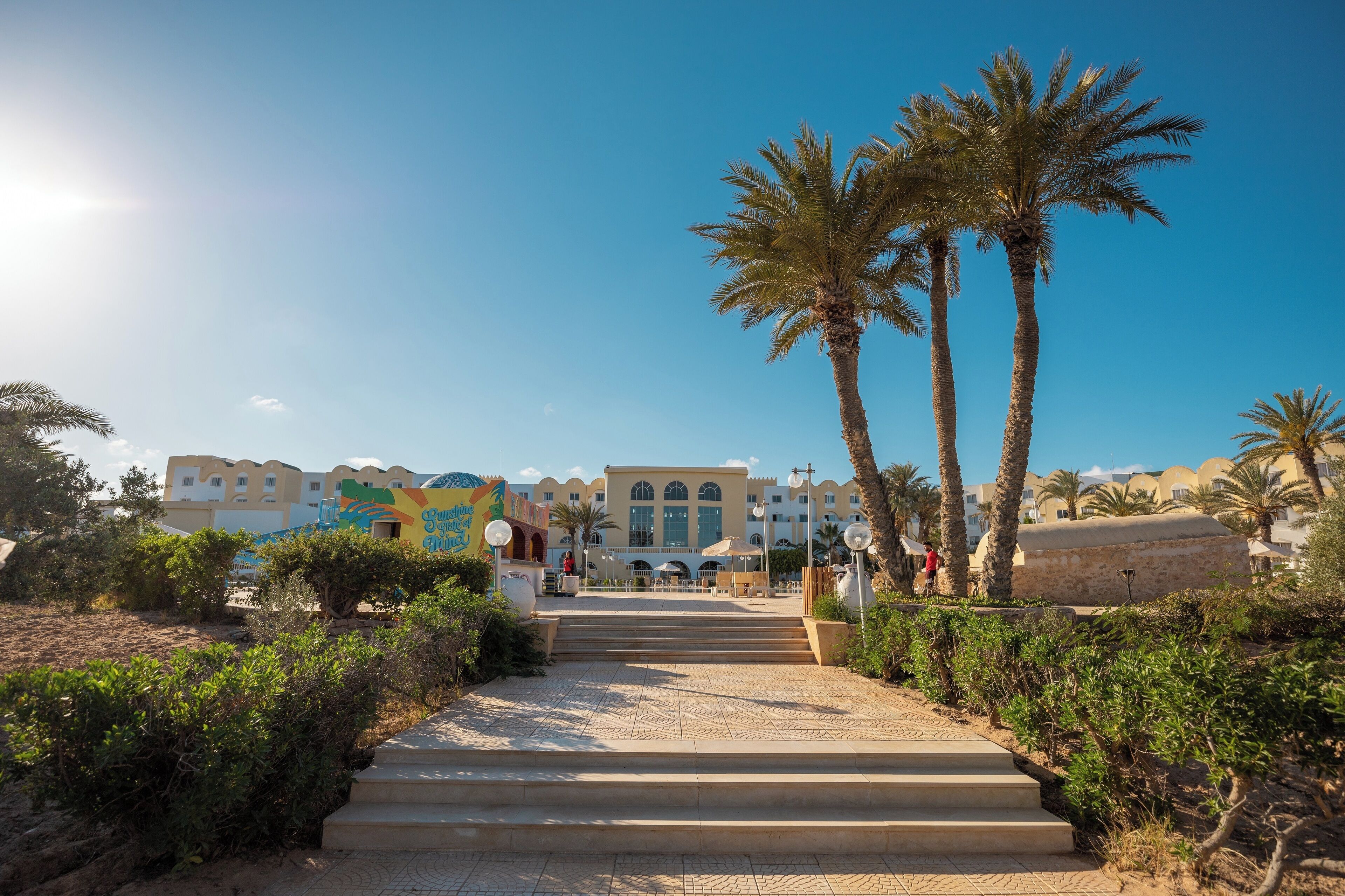 A hotel entrance with a paved pathway leading to a building, surrounded by palm trees and greenery.