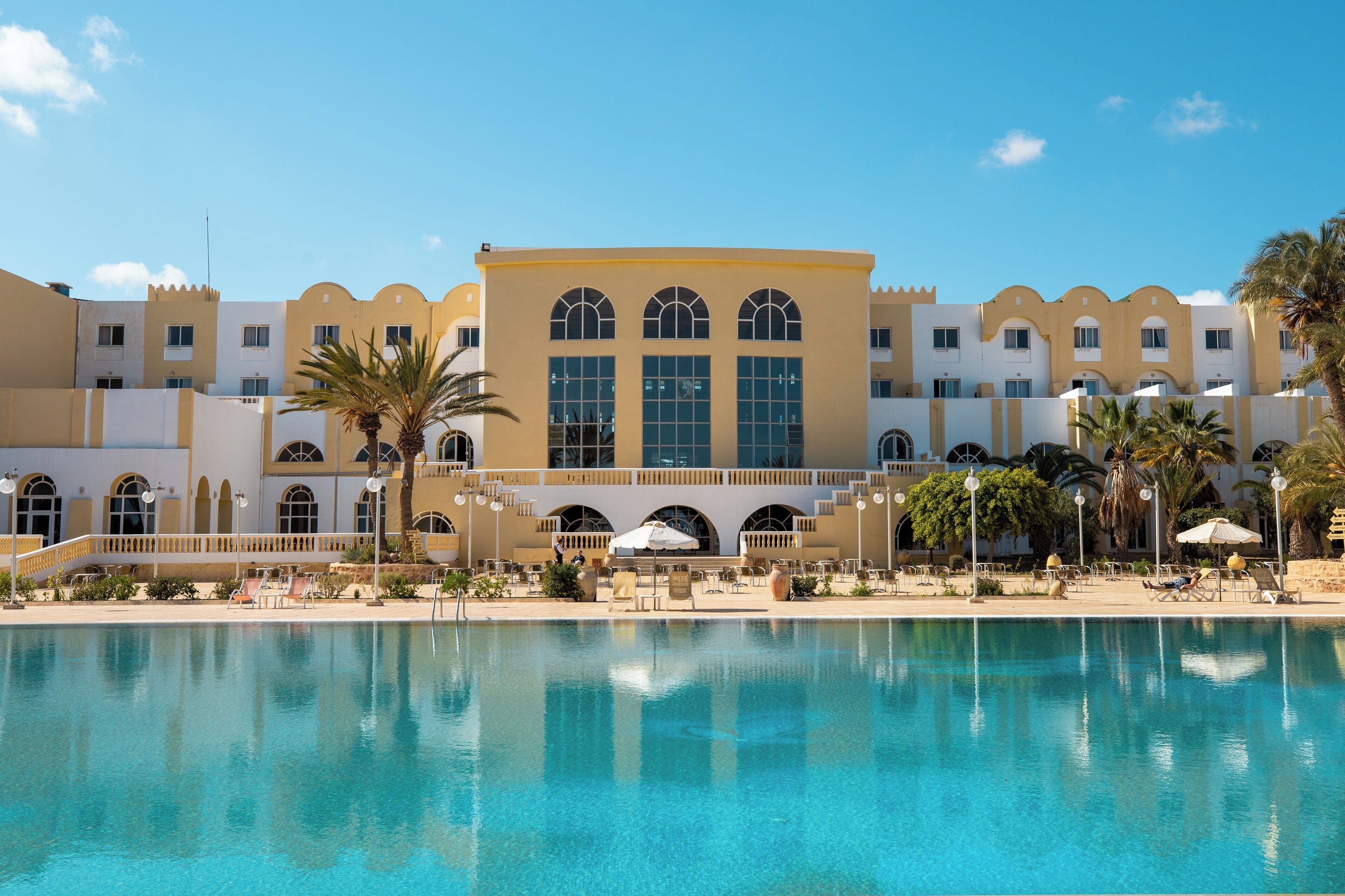 A hotel with a pool, palm trees, and a clear blue sky.