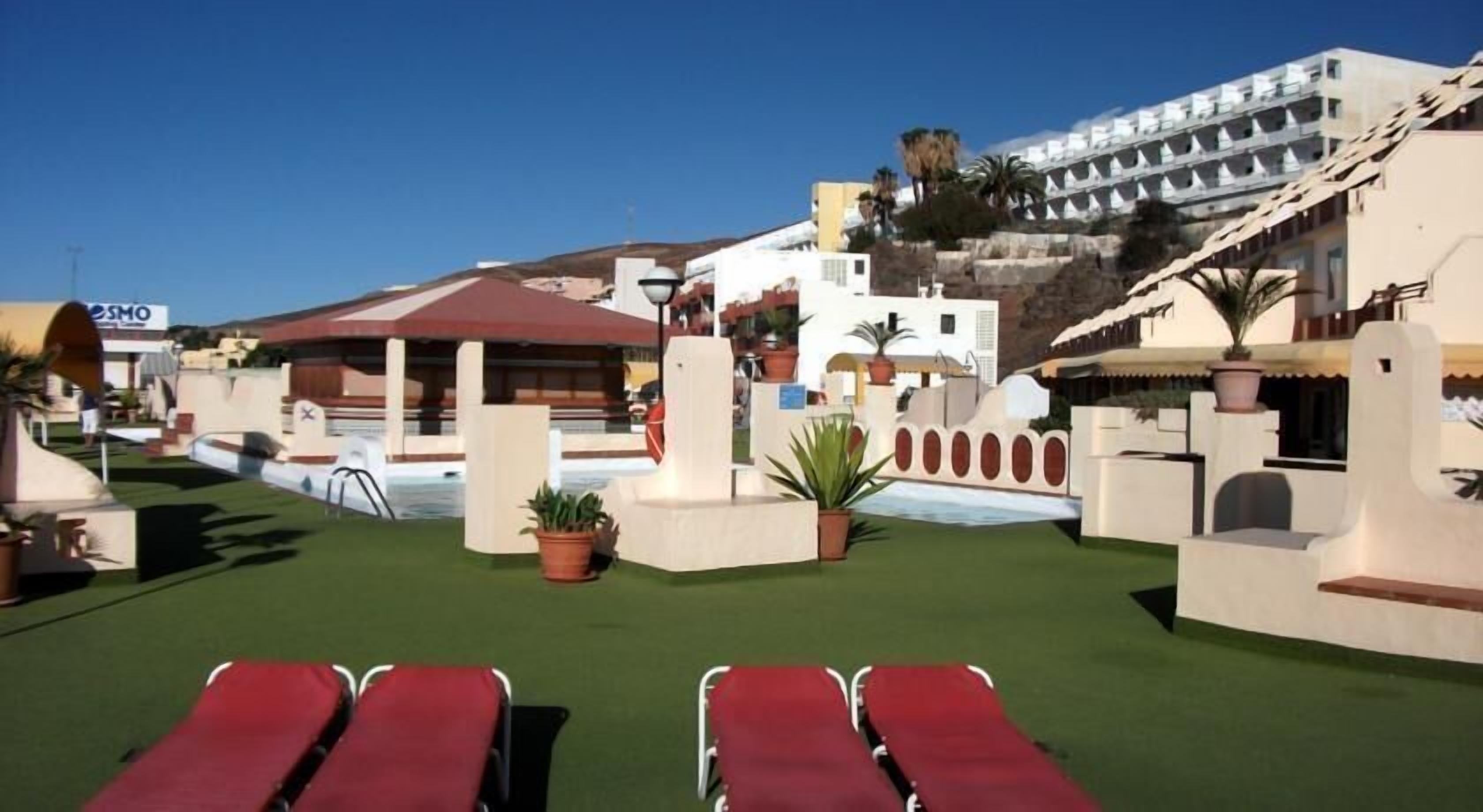 A pool area with red lounge chairs, a small garden with potted plants, and a building with balconies in the background.