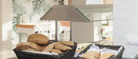 A breakfast buffet with various breads and pastries in wicker baskets, a glass-top table, and a modern interior with a palm leaf wallpaper.