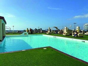 A swimming pool with a clear view of the ocean, surrounded by lounge chairs and shaded areas. - Palm Garden Apartments (Pajara)