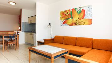 A modern living room with a wooden sofa, a dining table, and chairs. A kitchen is visible in the background.