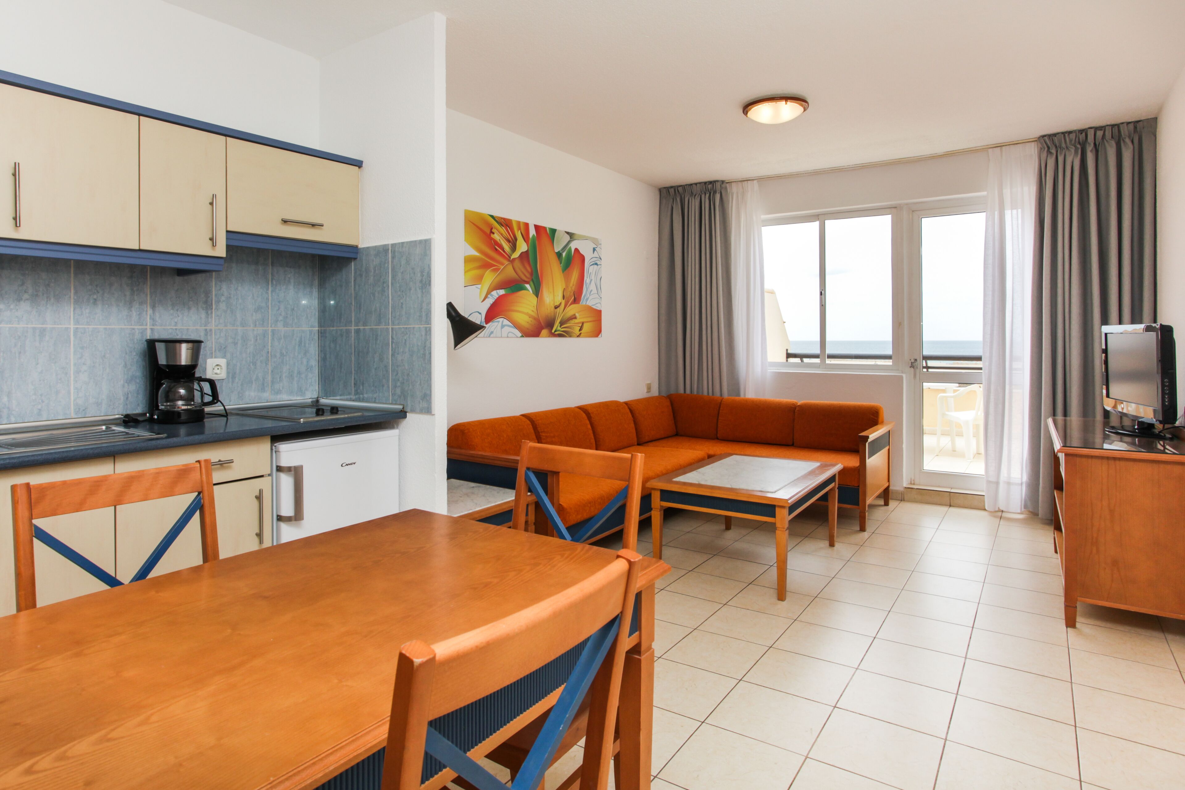 A modern kitchen with a coffee maker, a dining table with chairs, a sofa, and a TV on a wooden cabinet.