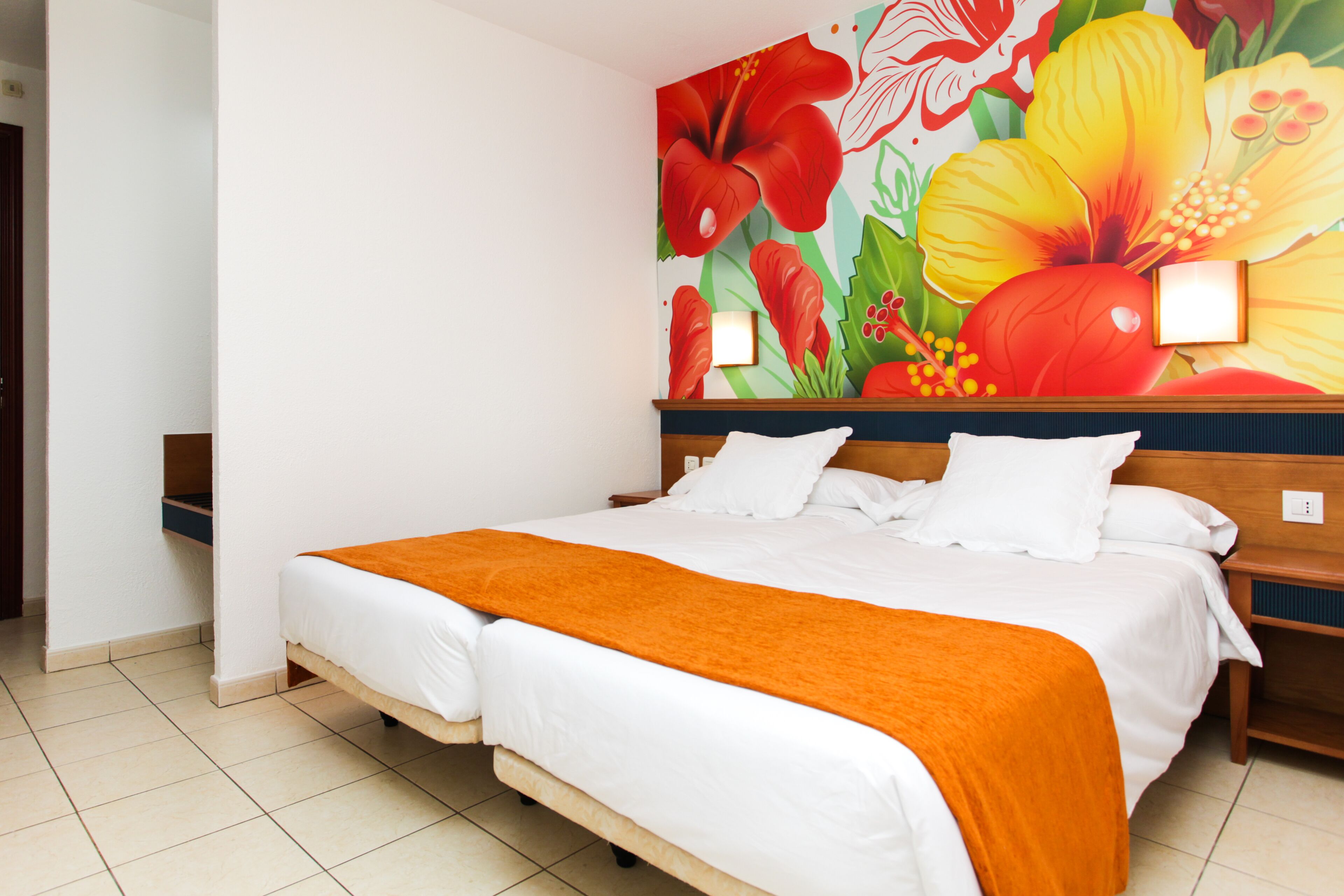 Habitación de hotel con cama, ropa de cama blanca, una manta naranja y un mural floral en la pared.