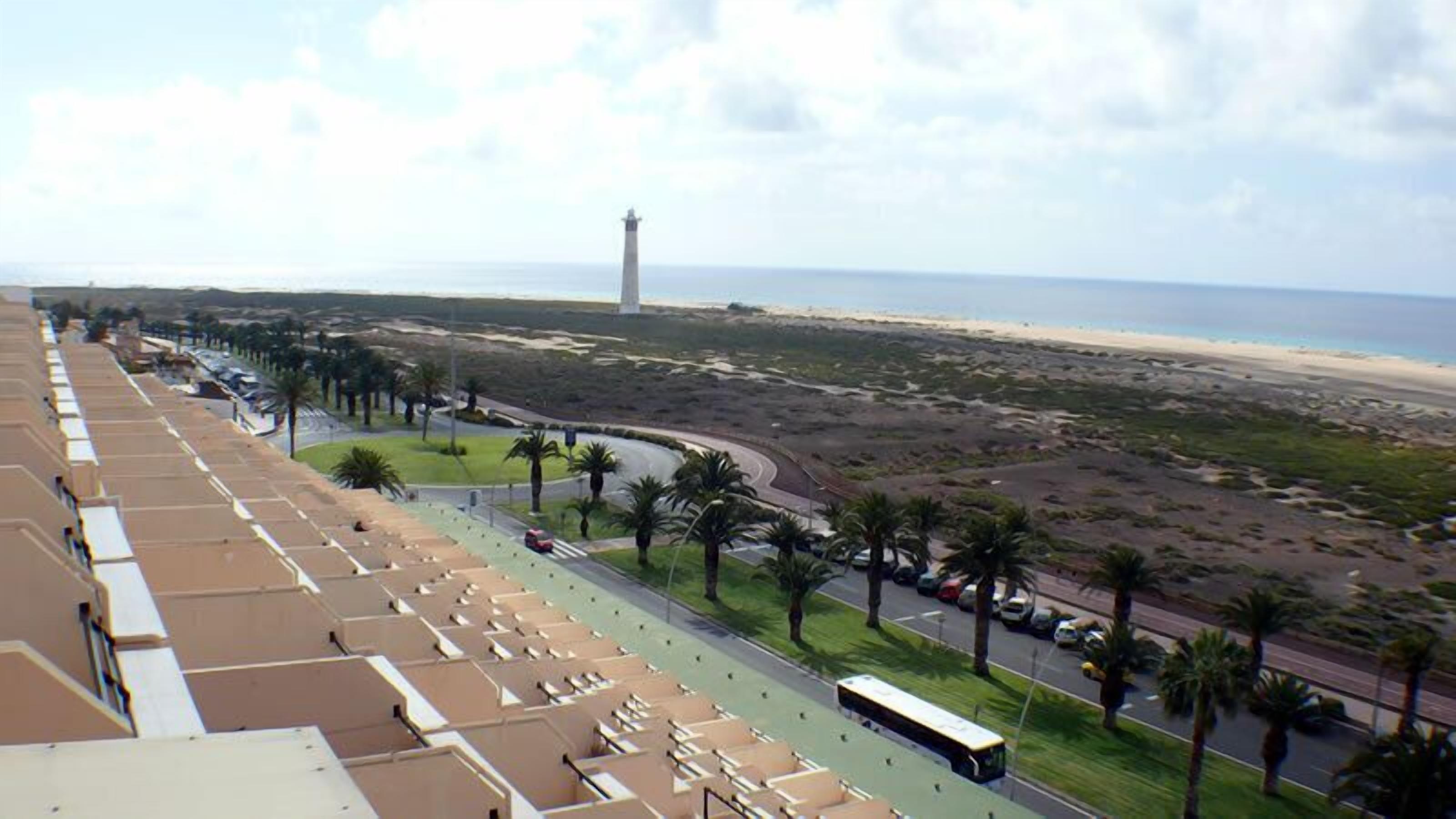 Une vue côtière comprenant une plage, un phare et une rangée de chambres d’hôtel donnant sur la mer.
