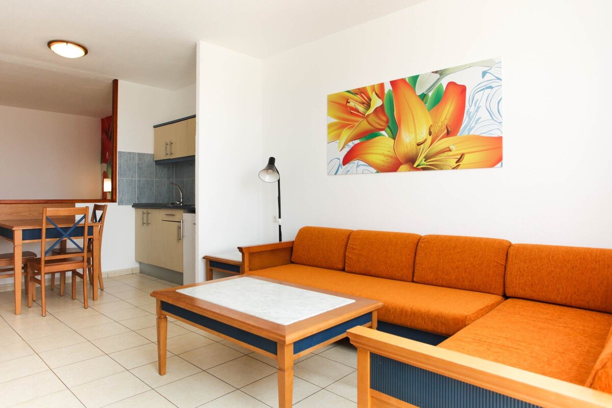 A modern living room with a sofa, a dining table with chairs, a kitchen area, and a painting on the wall.