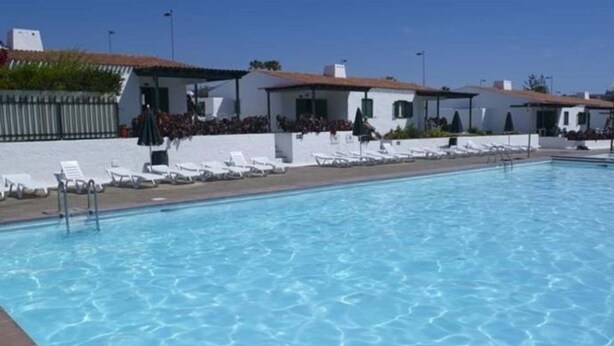 A swimming pool with lounge chairs and a white building with a tiled roof in the background. - Bungalows Las Tartanas III (San Bartolome de Tirajana)