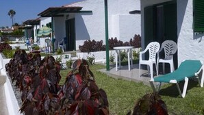 A white building with green shutters, a covered patio with white plastic chairs, and a row of red-leafed plants. - Bungalows Las Tartanas III (San Bartolome de Tirajana)