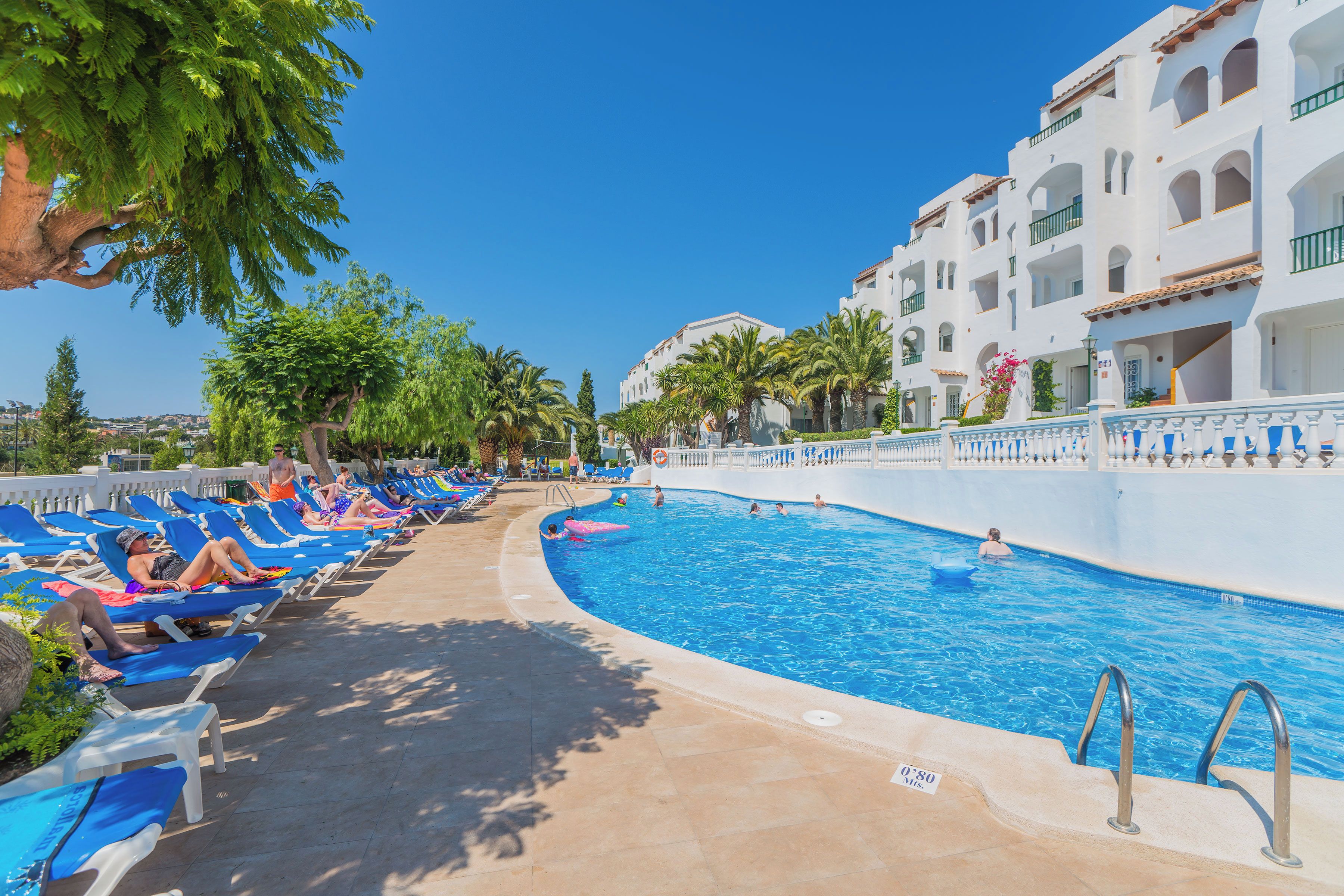 Un área junto a la piscina con sillas de descanso y una piscina, rodeada de edificios blancos con balcones.