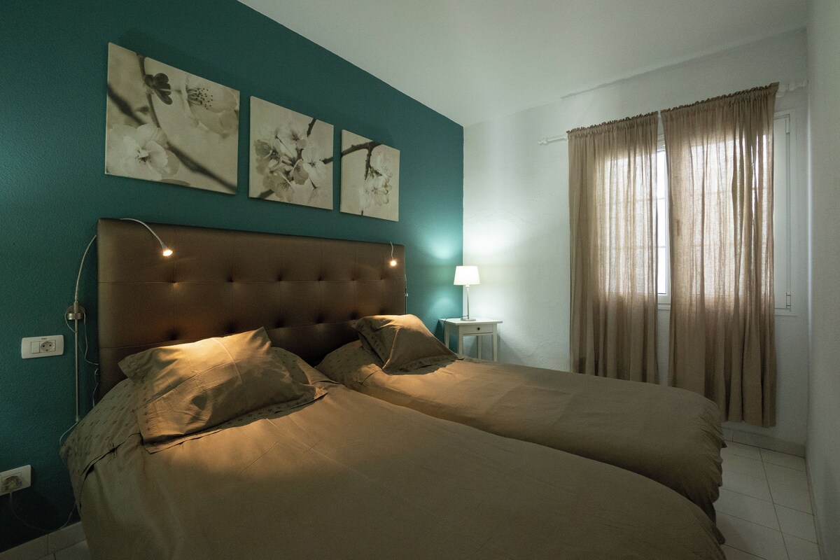 A bedroom with two beds, a headboard, a bedside table with a lamp, and a window with curtains.
