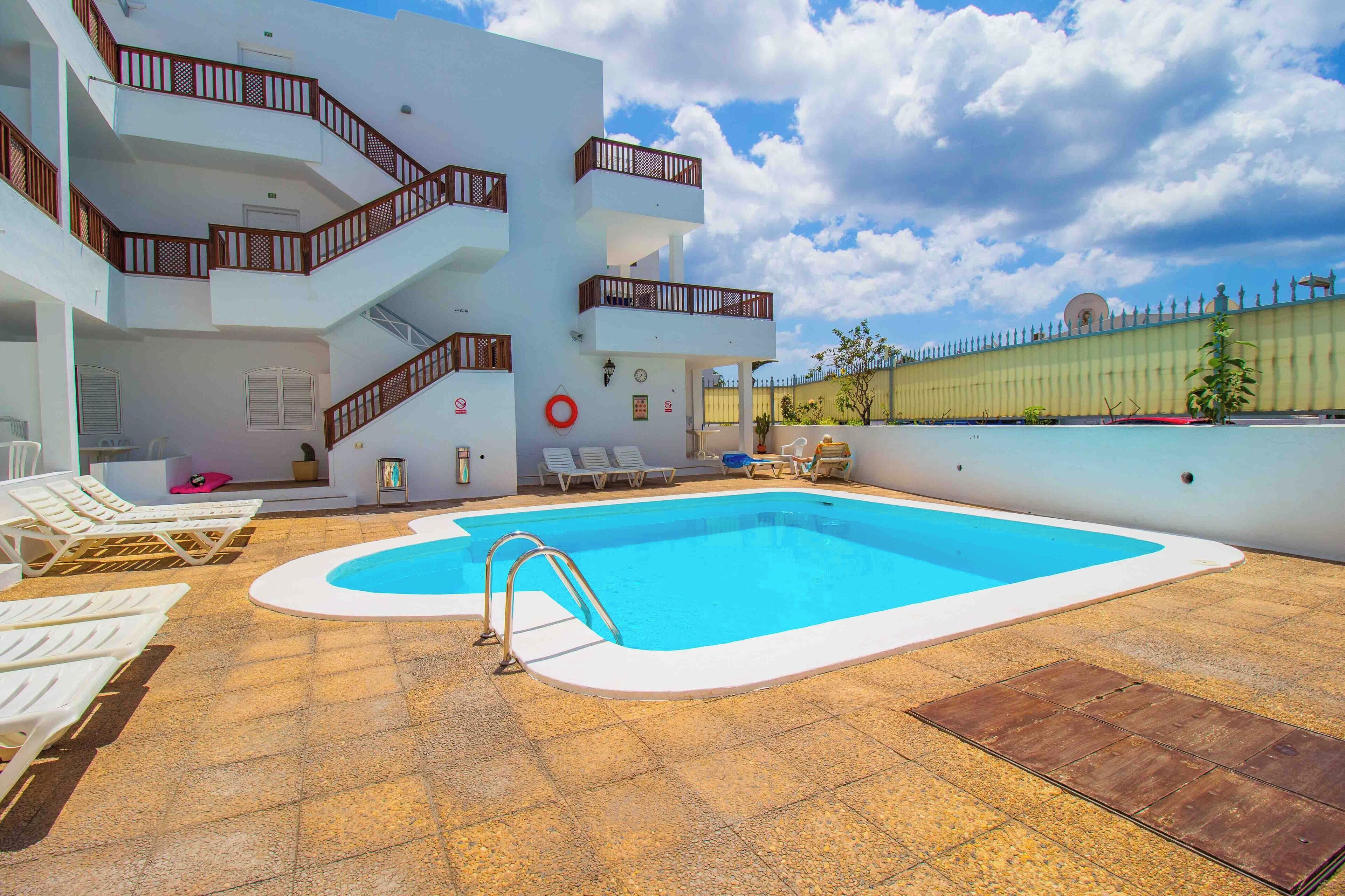 A pool area with a clear blue pool, white lounge chairs, and a multi-level building with balconies.