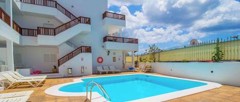 A pool area with a clear blue pool, white lounge chairs, and a multi-level building with balconies.