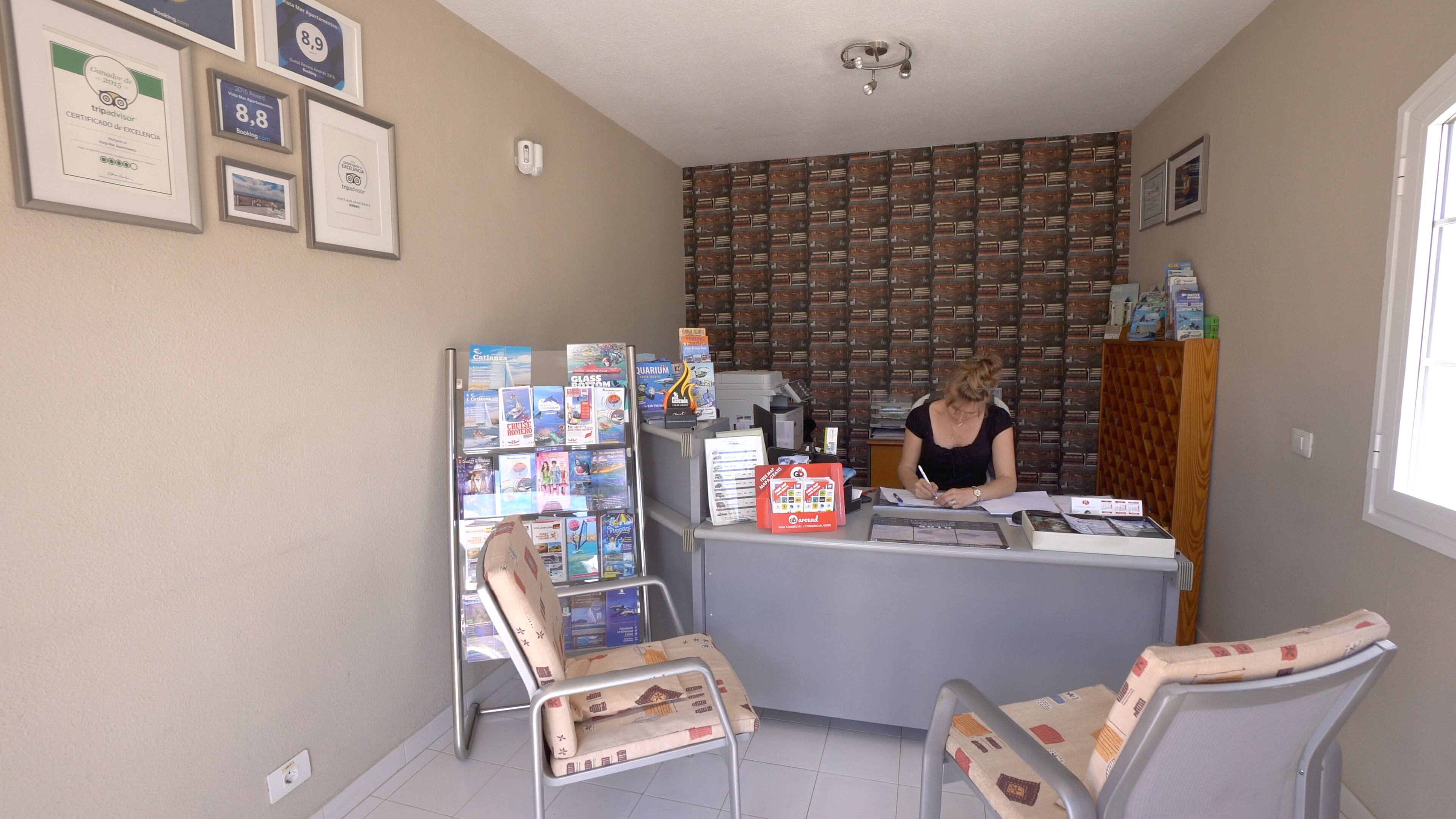Una reception con una donna che firma documenti, circondata da brochure di viaggio e una parete decorata.