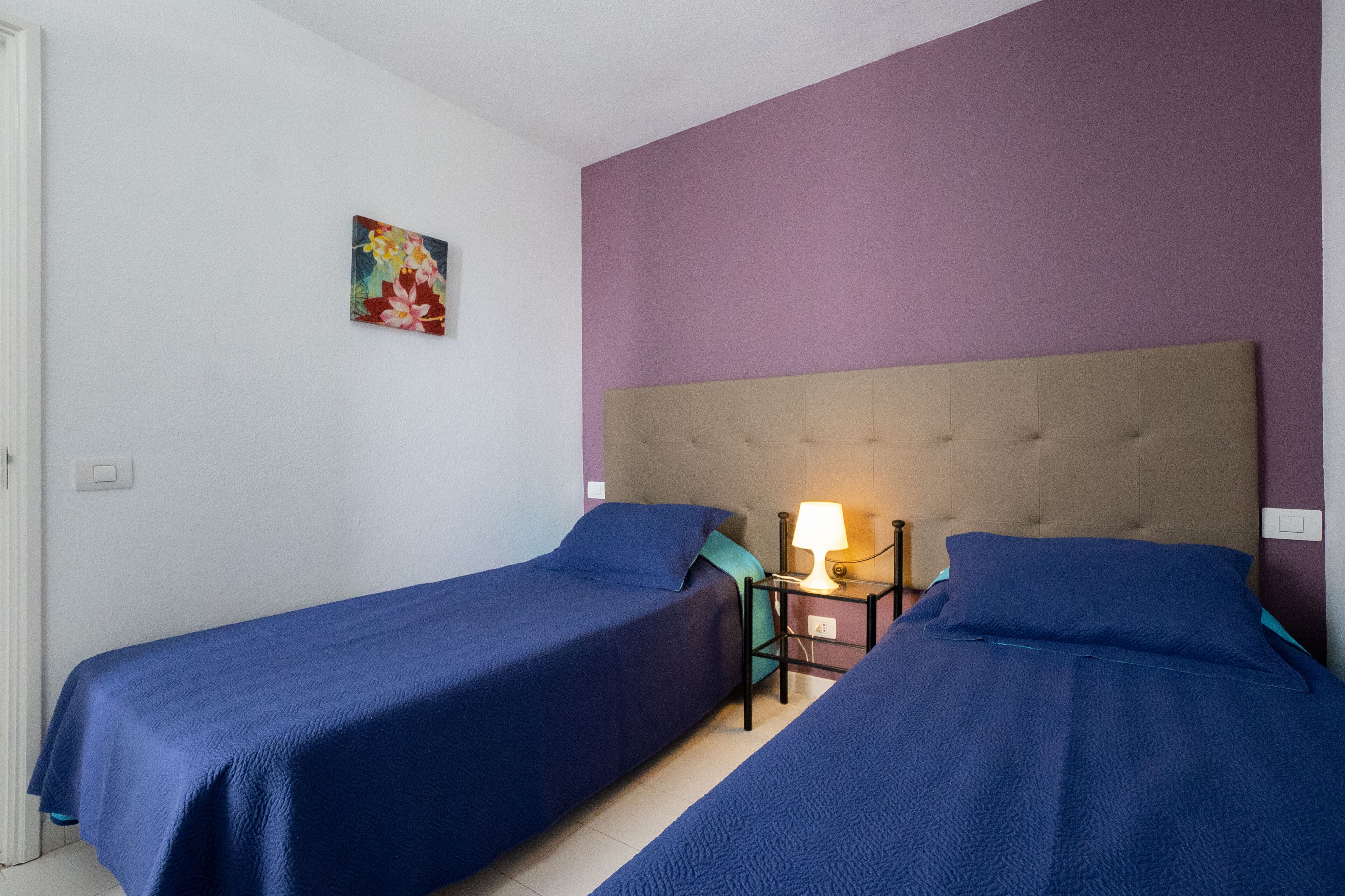 A room with two single beds, a nightstand with a lamp, and a small painting on the wall.